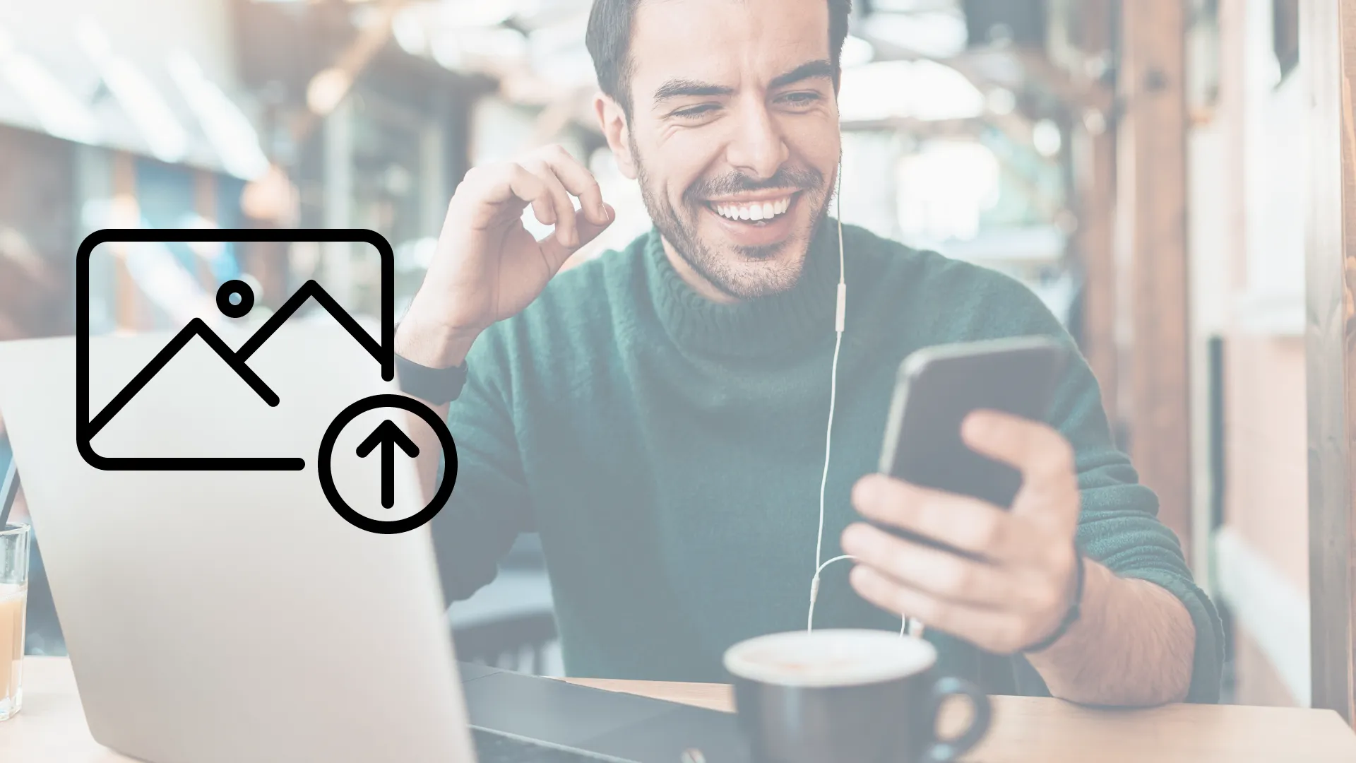 Hombre sentado en una mesa con smartphone en la mano, sonriendo frente a laptop